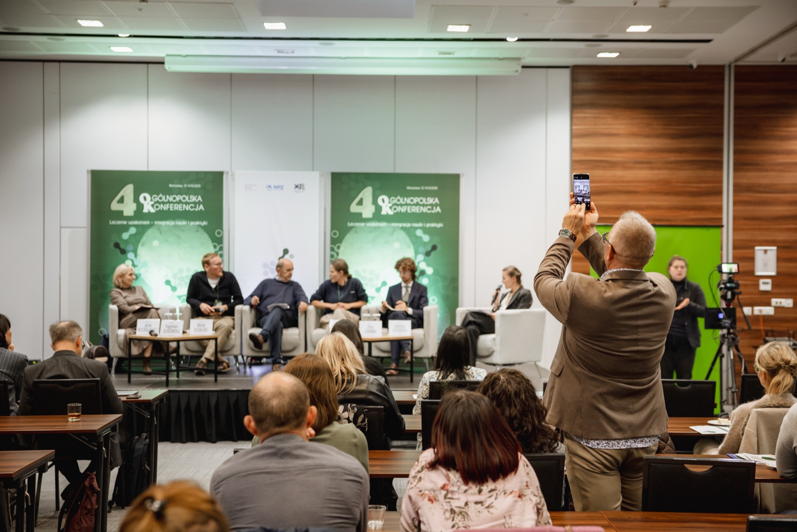 Sala konferencyjna z panelem na scenie, uczestnik fotografuje telefonem, w głębi kamera